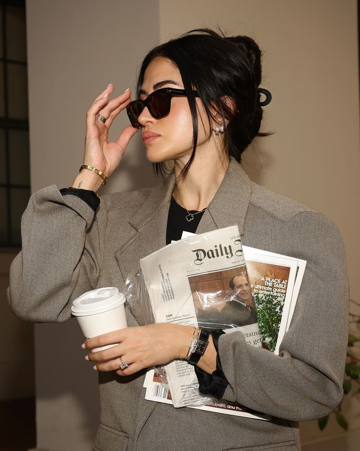 Stylish woman with sunglasses, holding coffee and newspapers, dressed in a gray blazer.