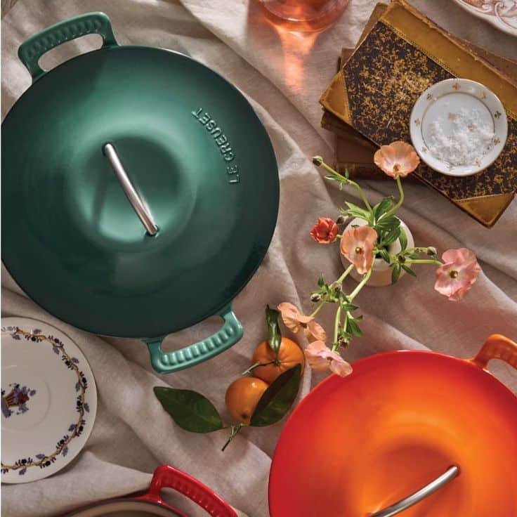Colorful Dutch ovens on a table with oranges, flowers, and plates, creating an inviting dining atmosphere.