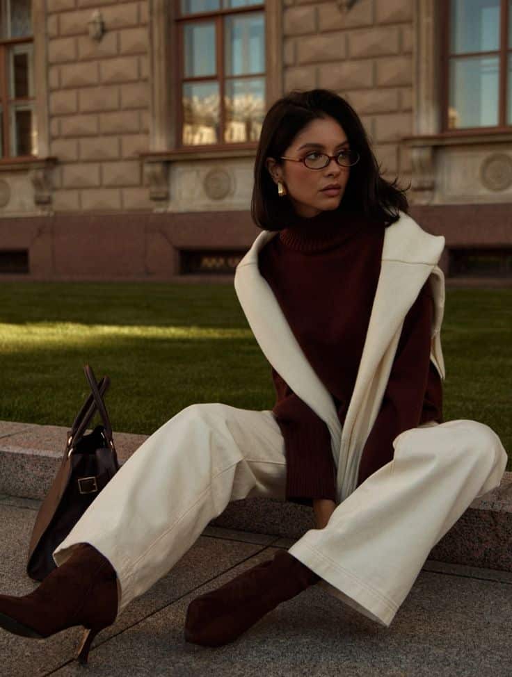 Stylish woman in a burgundy turtleneck and cream pants sitting outside a classic building, accessorized with glasses and boots.