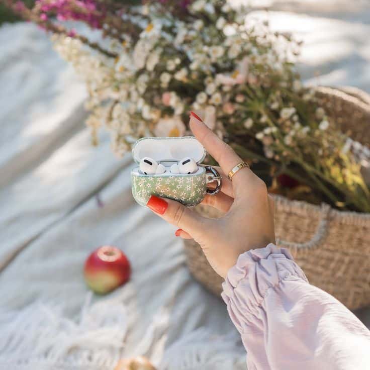 Hand holding floral AirPods case with earbuds against a picnic backdrop with flowers and apple.