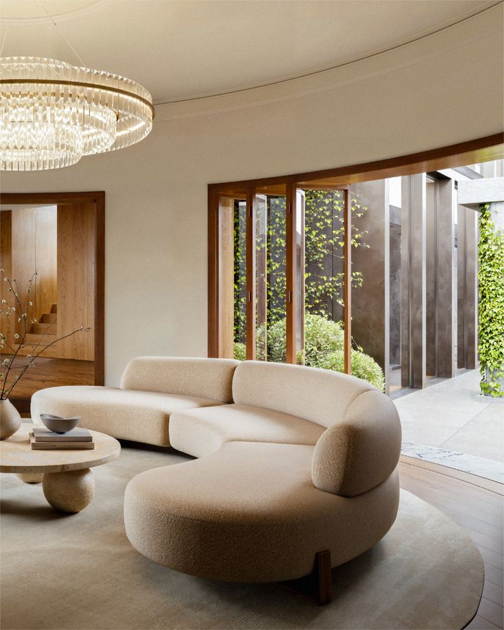 Modern living room with a curved beige sofa, elegant chandelier, and large windows opening to a garden view.