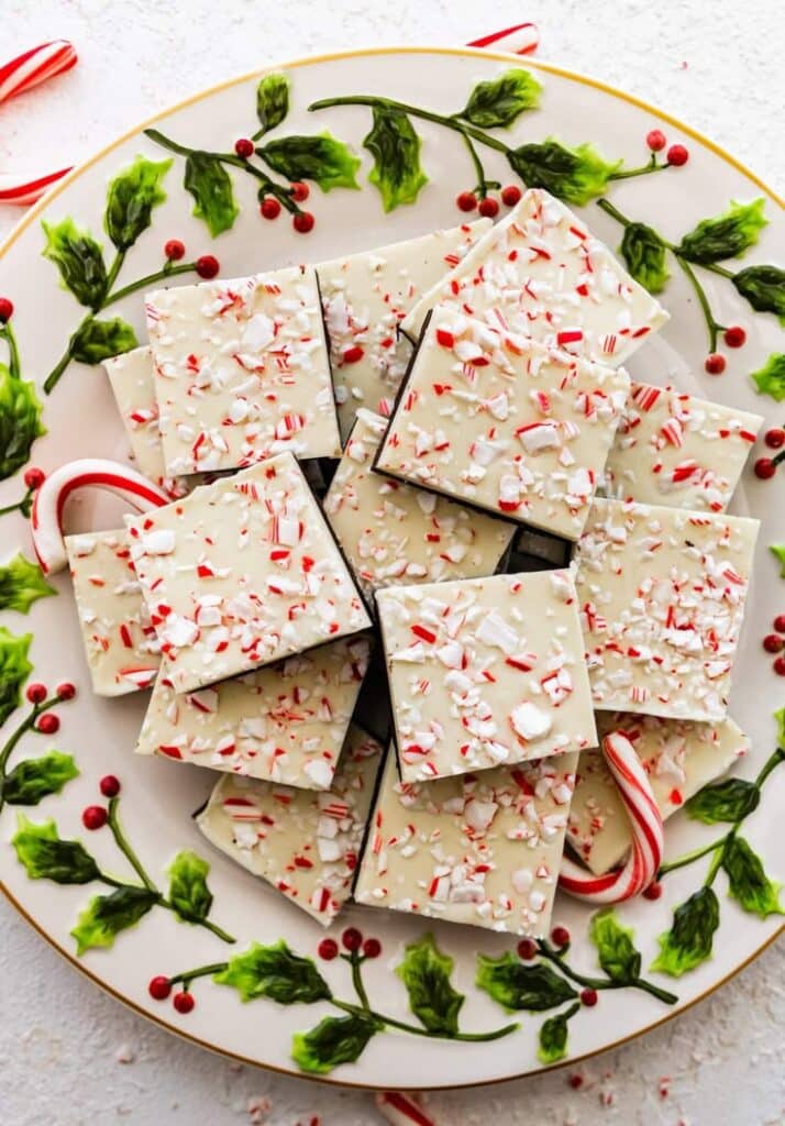 Festive peppermint bark squares on a holly-themed plate, topped with crushed candy canes. Perfect Christmas treat.