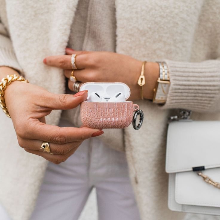 Woman holding AirPods in chic pink case, accessorizing with gold jewelry and a white handbag, showcasing elegant style.