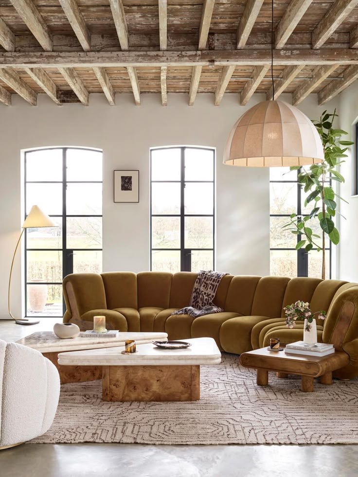 Stylish living room with brown sectional sofa, wooden tables, modern lighting, and large windows under rustic ceiling beams.