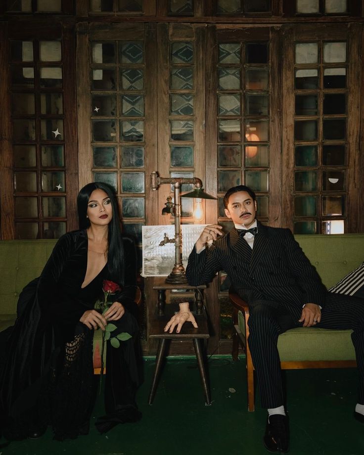 Couple in vintage attire sitting on a green sofa, with a mysterious hand sculpture and a rose, set in a classic wooden-paneled room.