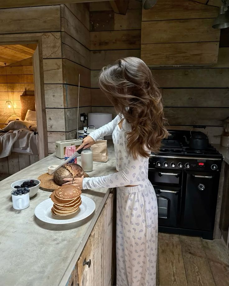 Woman preparing breakfast with pancakes, bread, and berries in a cozy wooden kitchen, creating a warm and inviting atmosphere.