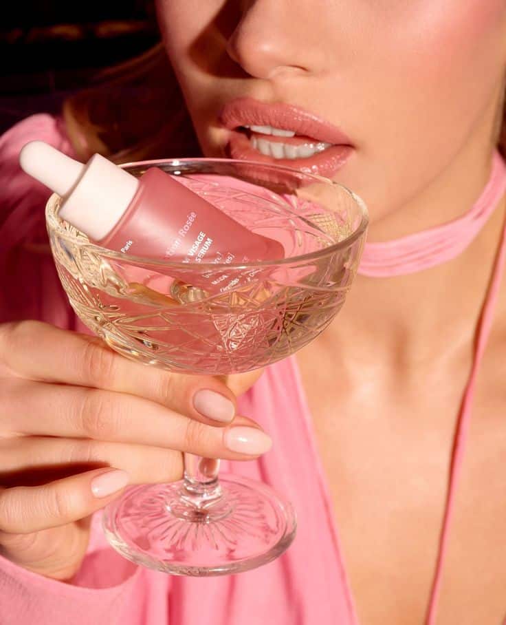 Woman holding elegant crystal glass with pink serum bottle, highlighting beauty and elegance. Perfect for luxury skincare ads.