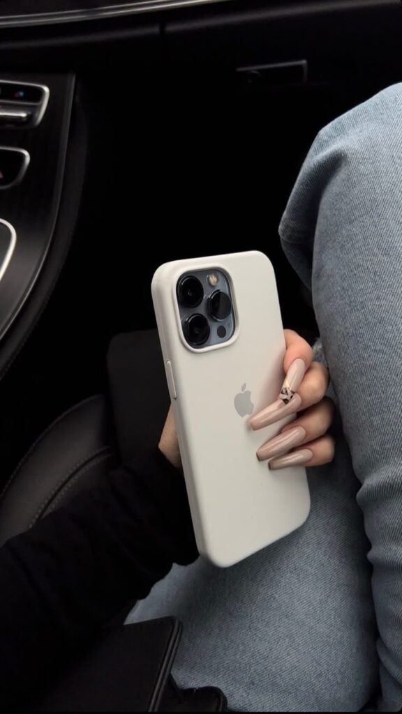 Person holding a white smartphone with a stylish case, seated with denim jeans, showcasing elegant manicured nails in a car.