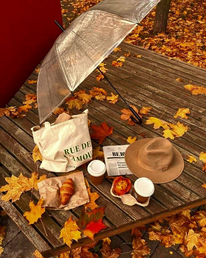 Autumn picnic on a wooden table with coffee, croissant, a hat, an umbrella, and fallen leaves. Cozy season vibes in the park.
