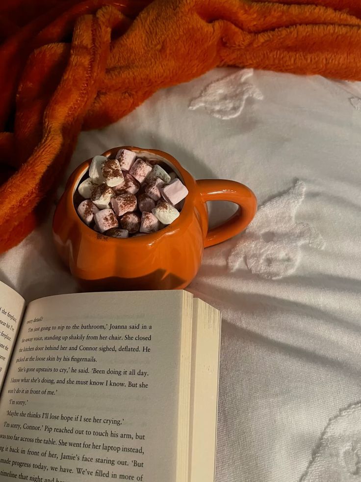 Cozy scene with a book and hot chocolate in an orange mug on a bed, topped with marshmallows, next to an orange blanket.