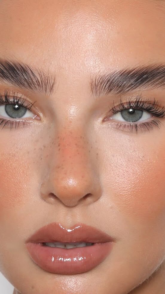 Close-up of a woman's face with natural makeup, glossy lips, bold brows, and highlighted skin, showcasing a fresh beauty look.
