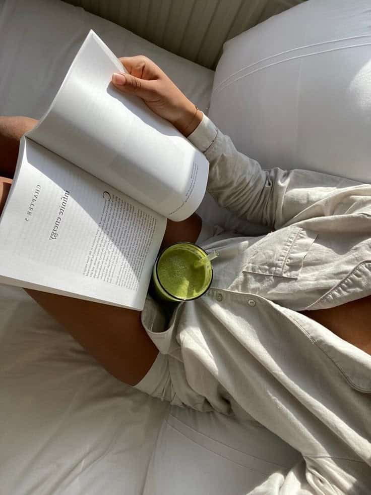 Person relaxing with a book and green smoothie in a cozy, sunlit bed, embracing a peaceful morning routine.