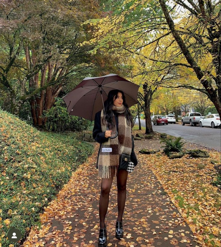 Woman with umbrella walking down a leaf-covered path in a cozy autumn setting, wearing a warm scarf and stylish outfit.