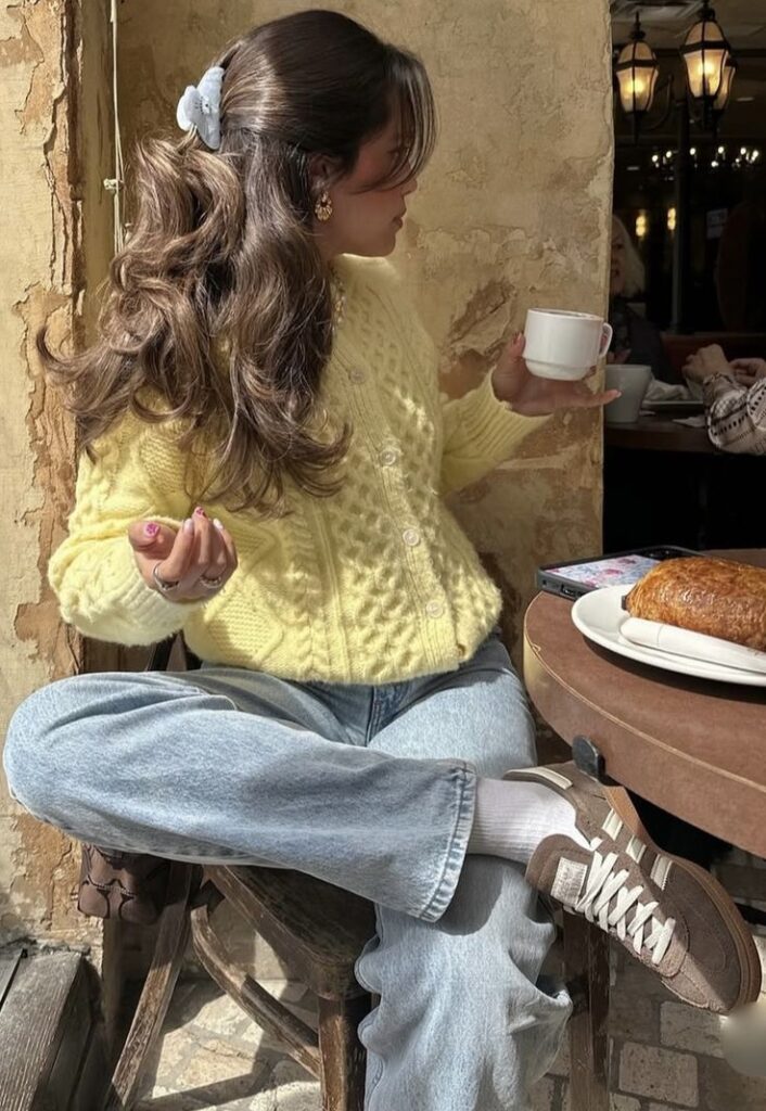 Woman in yellow sweater enjoying coffee and pastry at cafe, wearing jeans and brown sneakers. Cozy atmosphere, casual style.