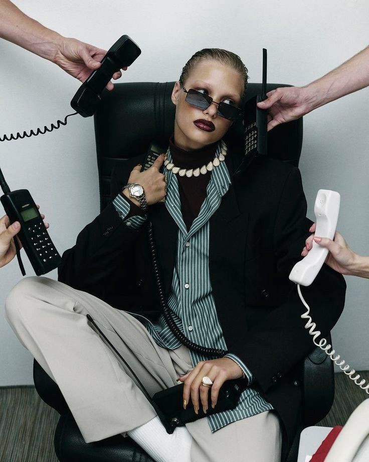 Stylish person in sunglasses surrounded by multiple retro phones, embodying a busy, fashionable professional look.