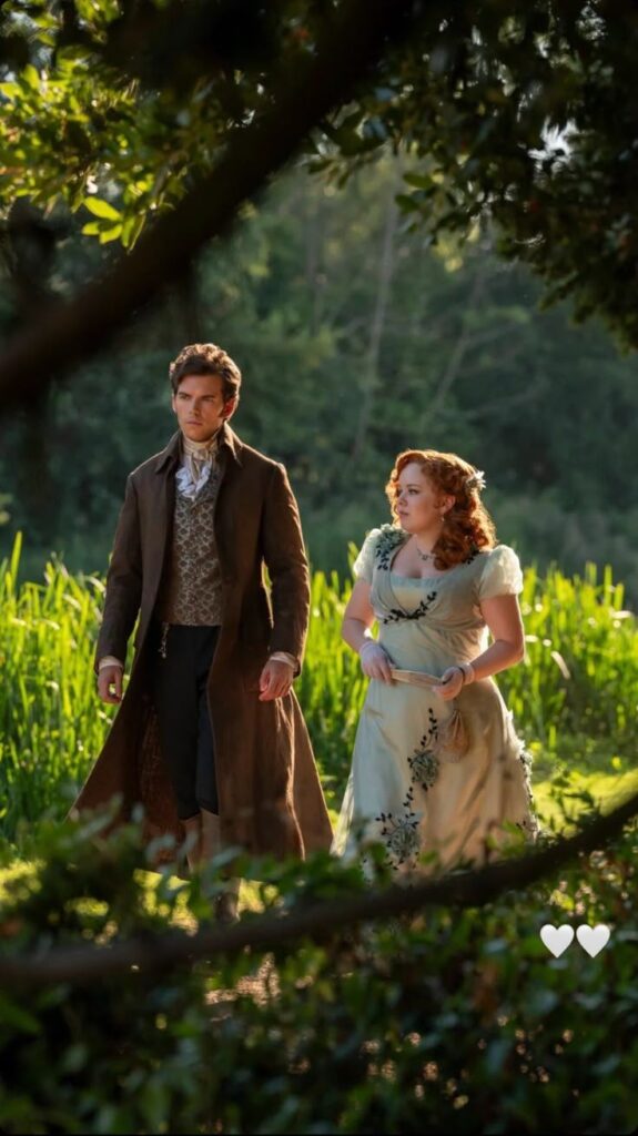 Two people in period attire strolling through a lush garden, surrounded by greenery and sunlight filtering through the trees.