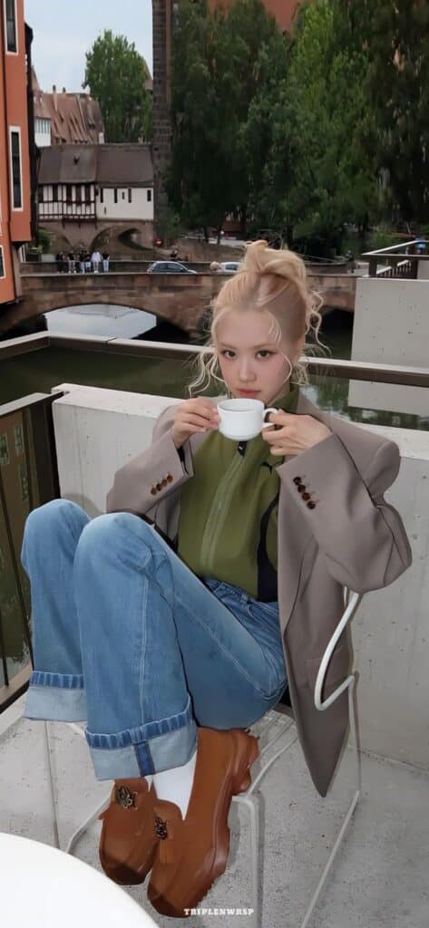 Person sipping coffee on a balcony, wearing a green shirt and blazer, with an urban riverside view in the background.