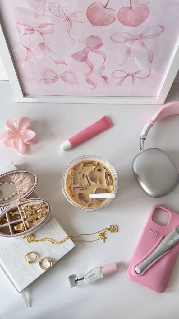 Flatlay of pink and gold themed accessories with iced coffee, headphones, beauty products, jewelry, and a pink poster.