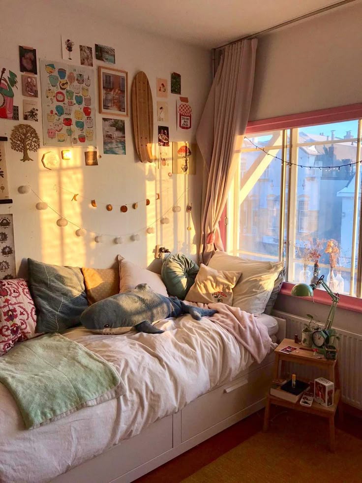 Cozy bedroom with wall art, string lights, pillows, and a plush toy. Sunlight streams through large windows with sheer curtains.