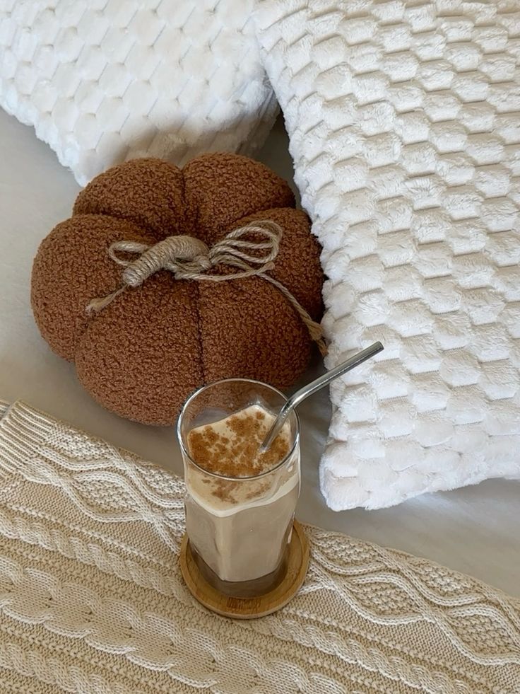 Cozy setting with a pumpkin pillow, textured white pillows, and a cinnamon latte in a glass with a metal straw.