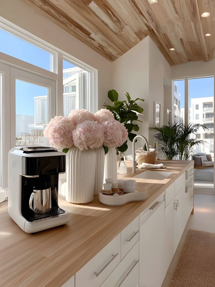 Modern kitchen with light wood countertops, coffee maker, and elegant vases of pink flowers, filled with natural sunlight.
