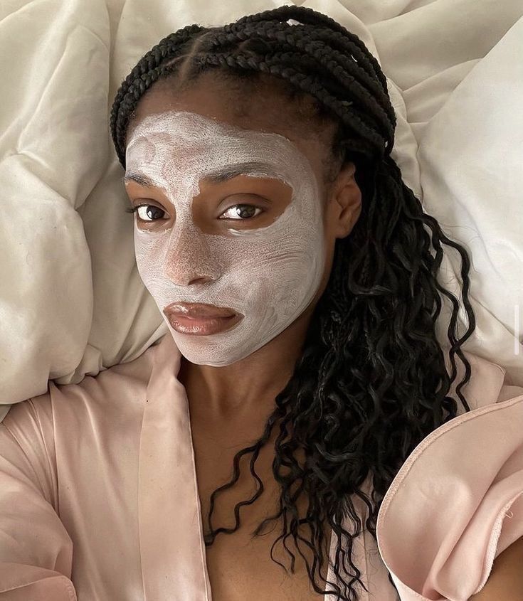 Woman relaxing with a clay face mask and braided hair, wearing a pink robe, lying on a bed. Skincare and relaxation concept.