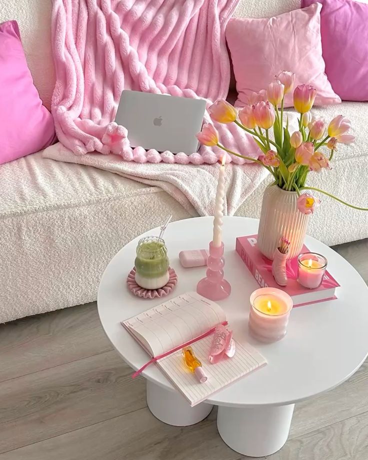 Cozy pink-themed living room with a laptop, flowers, candles, and notebook on a white table. Feminine workspace vibes.