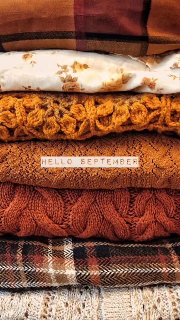 Stack of cozy autumn sweaters and blankets with Hello September message, highlighting warm fall colors and textures.