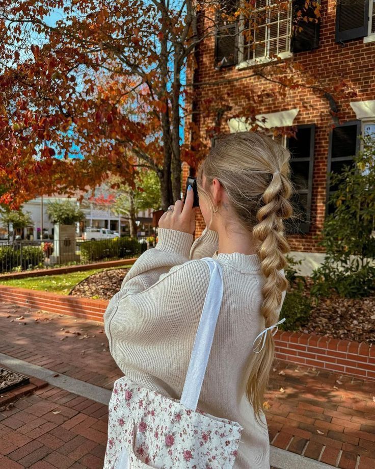 Woman with a braid takes a photo of colorful autumn leaves on a tree in a quaint brick street setting.