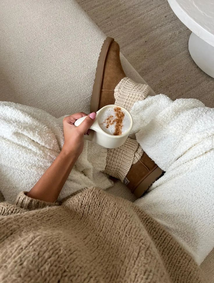Cozy scene with a person in wool socks and brown slippers holding a cinnamon latte on a plush beige couch.