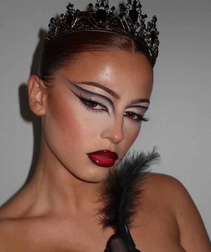 Elegant woman with dramatic makeup, wearing a black crown and holding a black feather against a neutral background.
