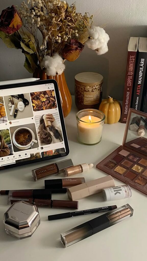 Cozy autumn vibe with makeup, tablet showing fall images, candle, and books on a desk. Perfect ambiance for beauty lovers.