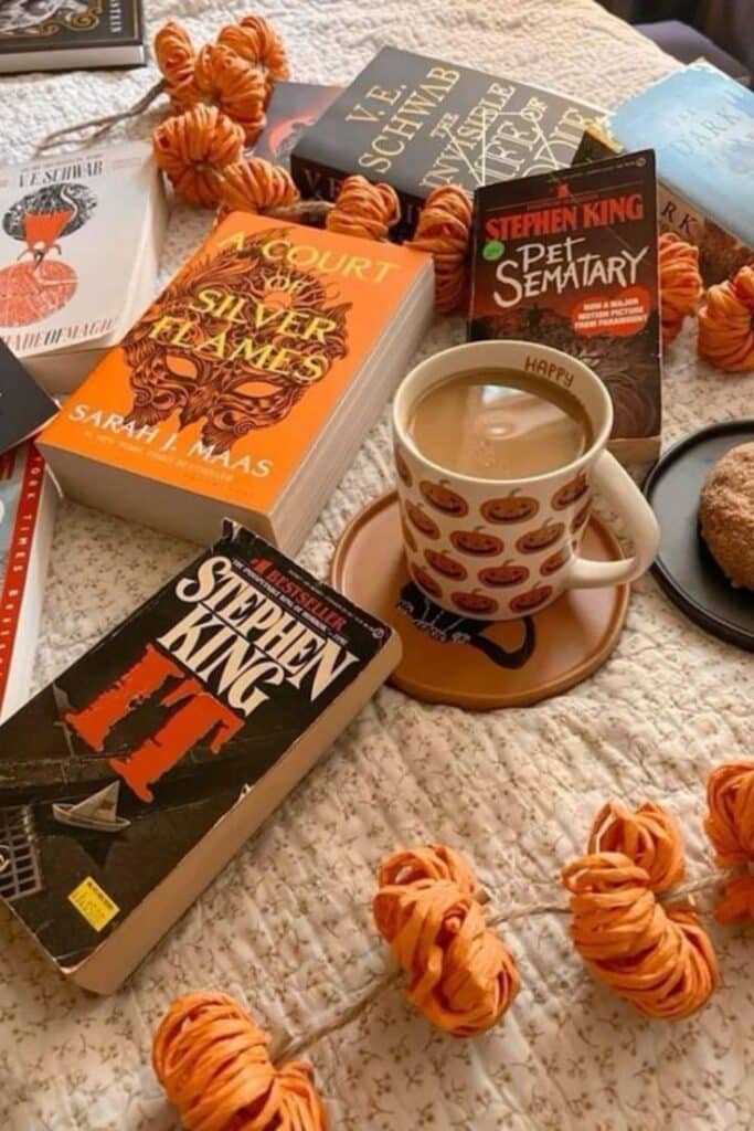Cozy autumn reading setup with popular books, pumpkin-themed mug of coffee, and orange decor on a quilted blanket.