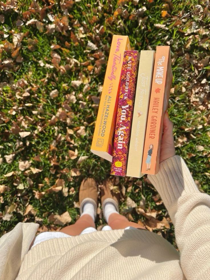 Person holding four colorful books on a sunny day, standing on grass with autumn leaves. Cozy vibe with tan sweater and shoes.