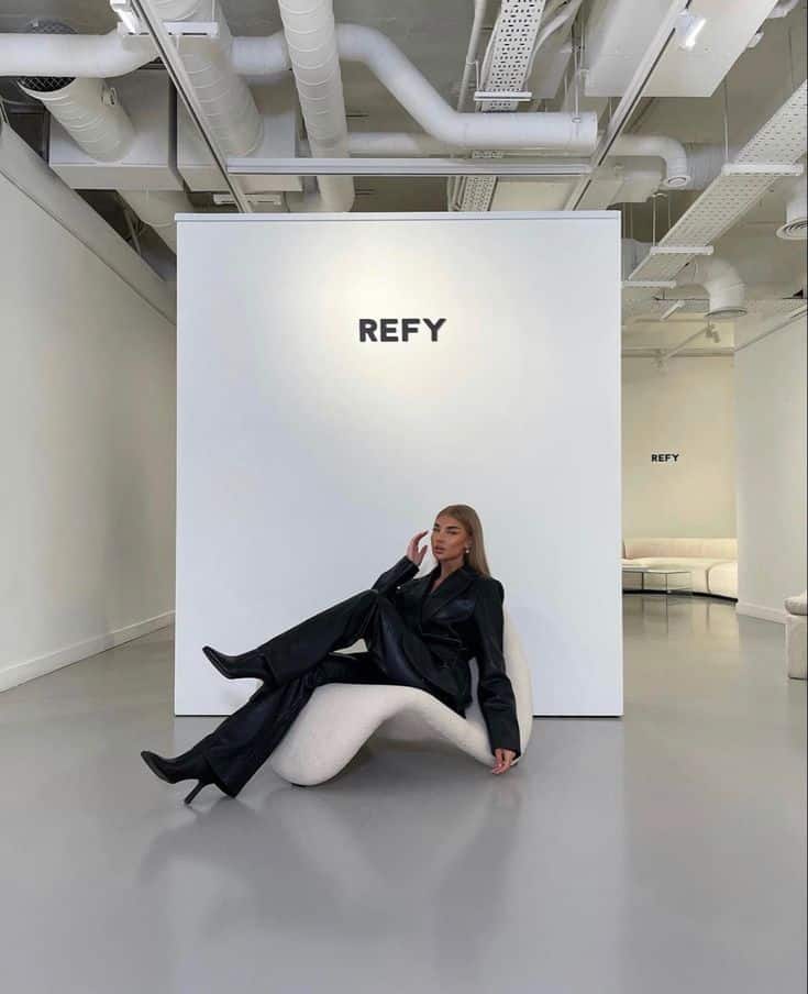 Person in black leather suit sitting on modern chair at REFY showroom with minimalist design and industrial pipes overhead.