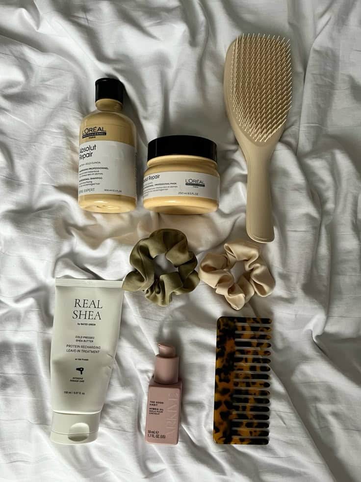 Haircare essentials: shampoo, mask, brush, scrunchies, shea treatment, serum, and comb on a white fabric background.