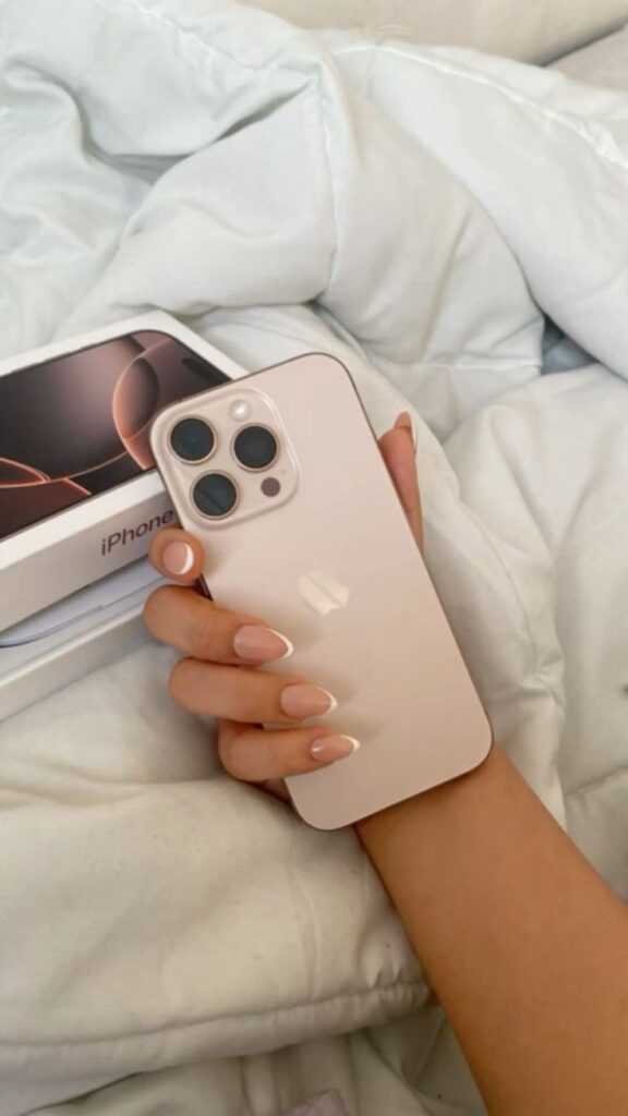 Hand holding a new iPhone with a box, on a bed with white sheets. Modern smartphone photography setup.
