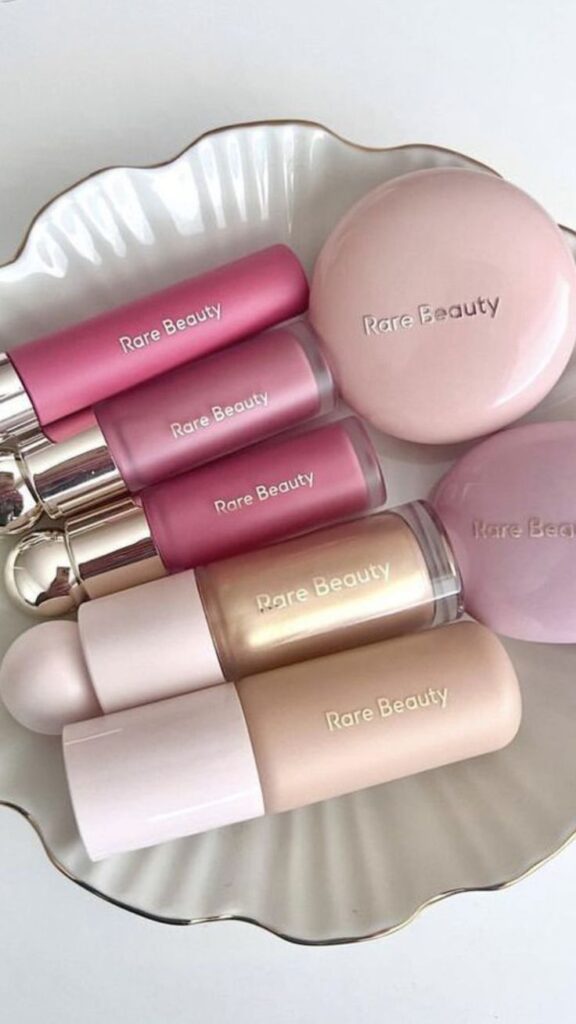 Assorted Rare Beauty makeup products arranged on a decorative plate, featuring blushes, lipsticks, and compacts in soft pinks and neutrals.
