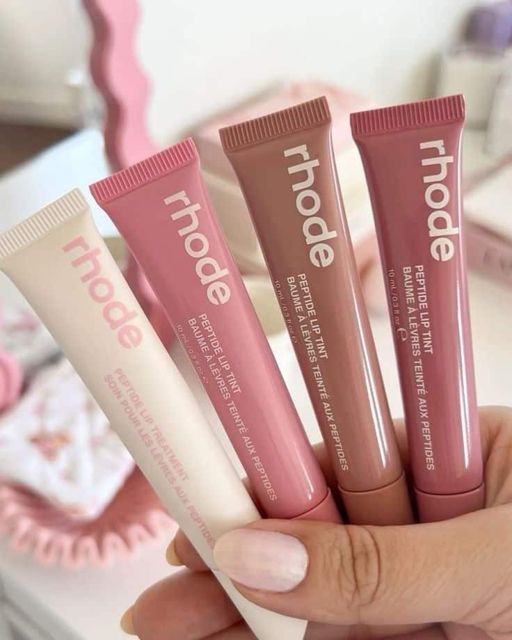 Four Rhode peptide lip tint tubes in various shades, held in hand, showcasing skincare cosmetics for glowing lips.