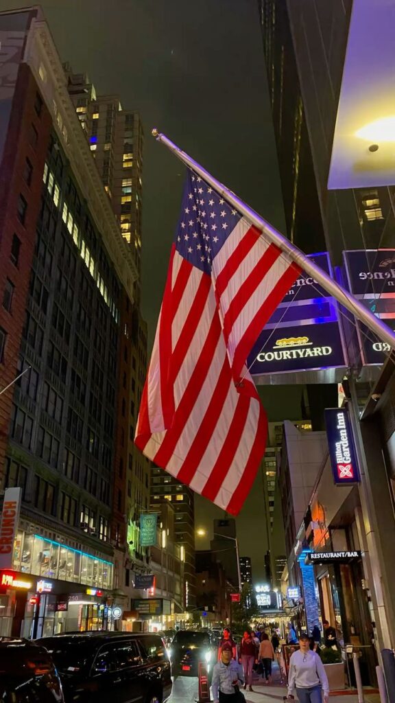 American flag on a bustling city street at night, with vibrant building lights illuminating the scene.