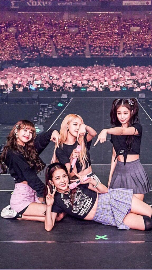 Four performers posing energetically on stage in front of a cheering crowd at a concert venue.