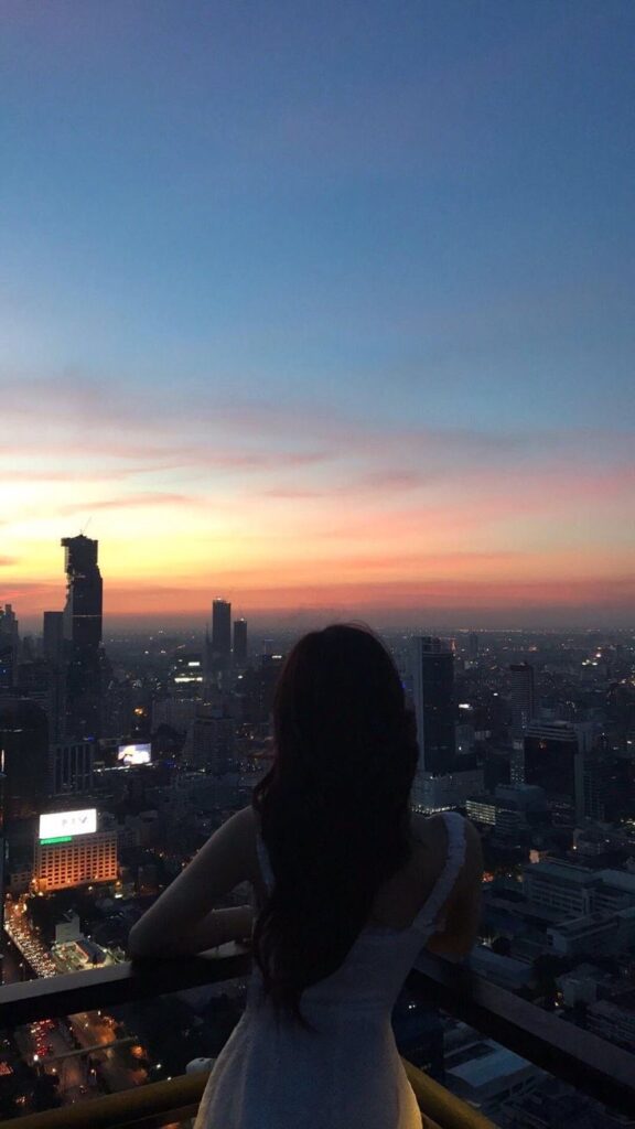 Woman admiring a vibrant cityscape at sunset from a high vantage point, with multi-colored sky above urban skyline.