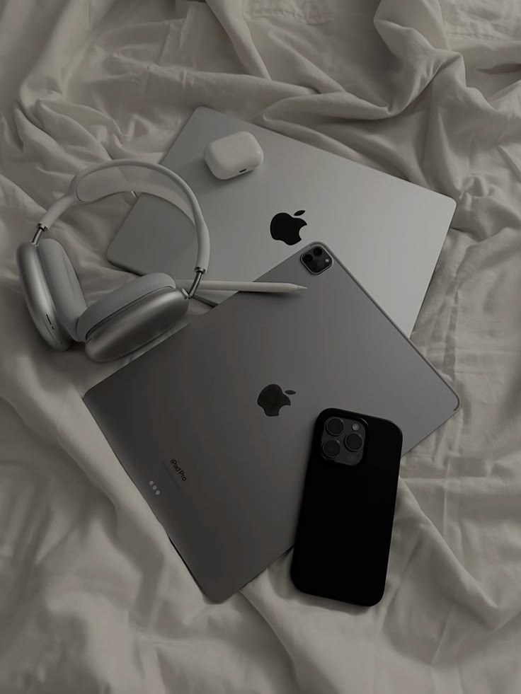 Apple devices on bed: MacBook, iPad, iPhone, AirPods, and headphones. Minimalist tech setup in neutral tones.