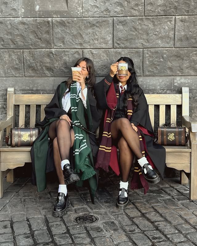 Two people in wizard robes sit on a bench, sipping butterbeer, with Hogwarts-themed chests beside them.