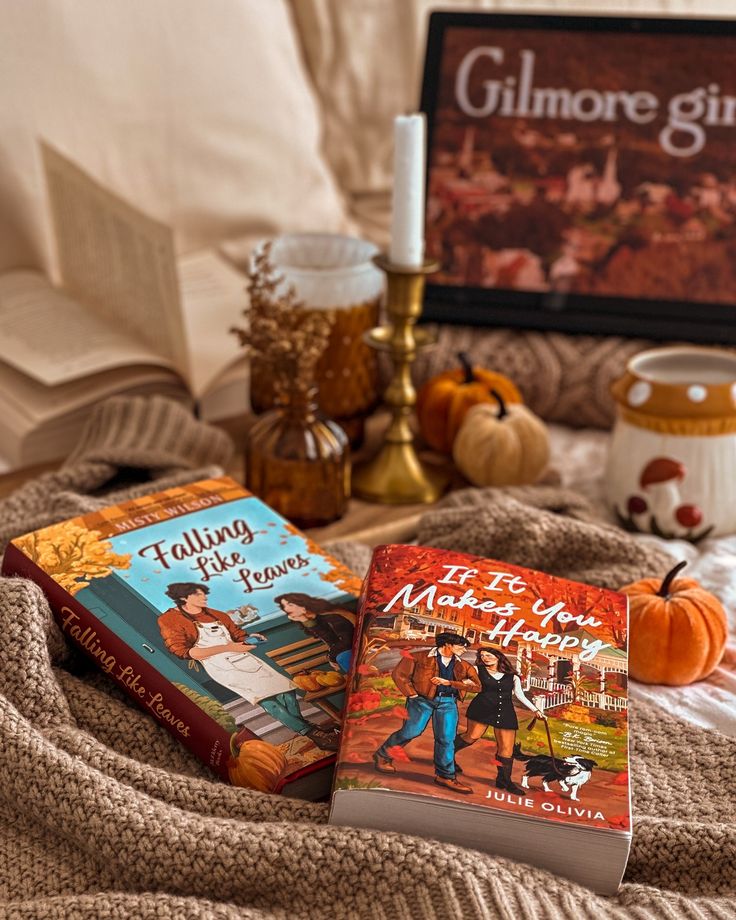 Cozy fall scene with two autumn-themed books, pumpkins, a knit blanket, and candles on a soft bedspread.