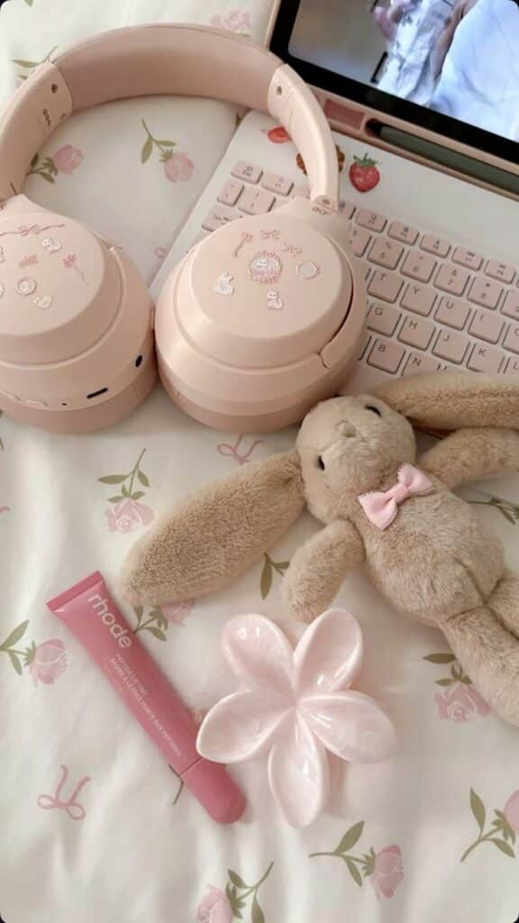Cute pink workspace with headphones, plush bunny, lip gloss, flower dish, and laptop on floral bedspread. Cozy aesthetic vibes.