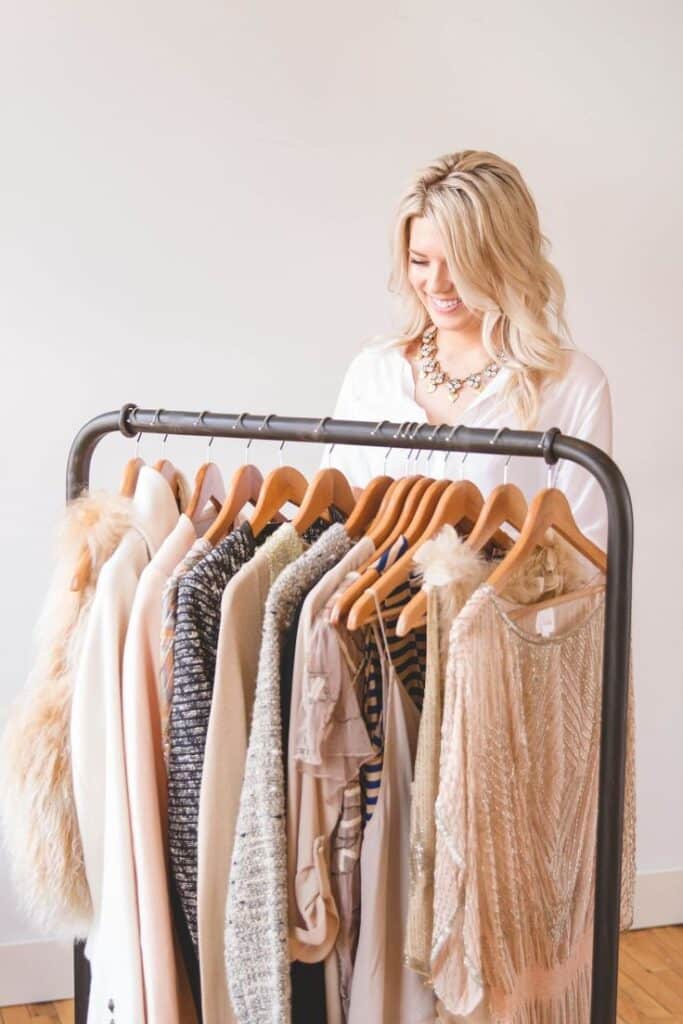 Blonde woman smiling while looking at a clothing rack with stylish outfits and hangers in a bright room. Fashion and style concept.