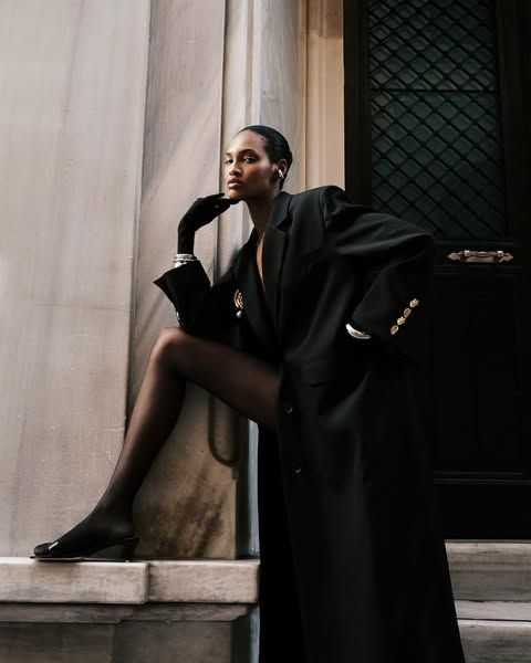 Fashionable woman in a black coat poses against a marble wall, exuding elegance and confidence.