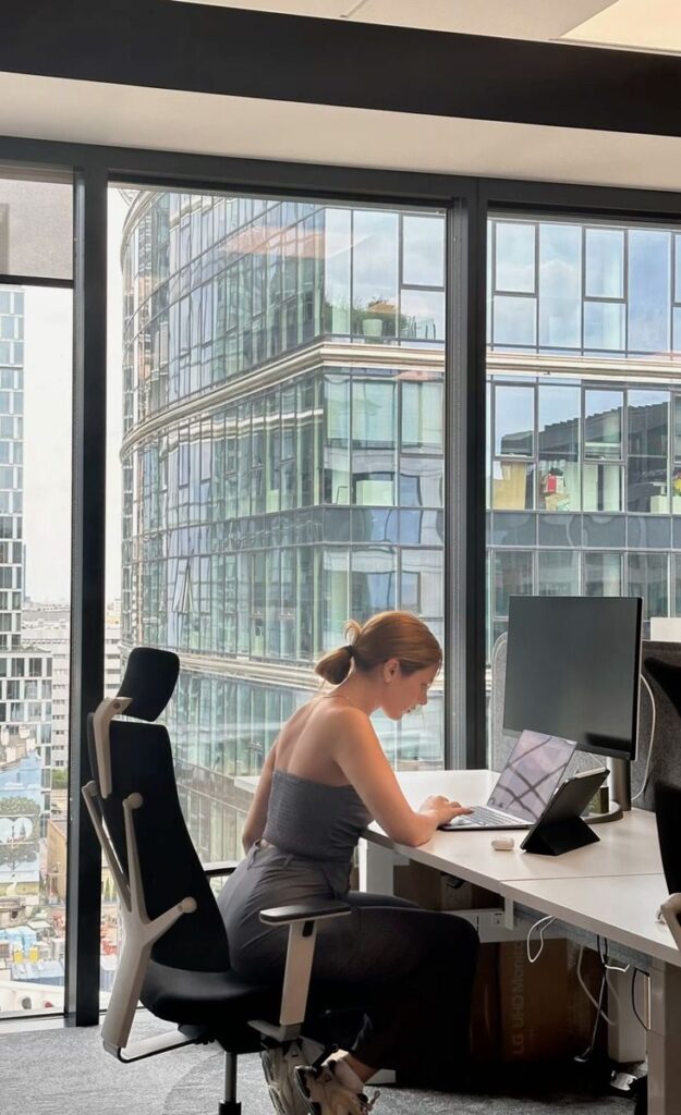 Woman works on laptop in modern office with cityscape view through large windows.