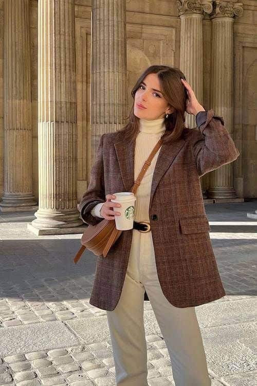 Stylish woman in a brown blazer holds a coffee cup outside historic building with columns, showcasing chic urban fashion.